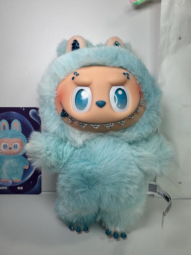May include: A plush toy in the shape of a creature with a light blue furry body and head covering. The toy has large blue eyes, a smiling mouth with braces, and small ears. A small card with a similar character is visible.