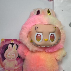 May include: A plush toy in shades of pink, yellow, and orange, resembling a bunny with a hood. The toy has a detailed face with large eyes and a small yellow nose. A card with a matching illustration is next to the toy. The word "LOYALTY" is printed on the card.