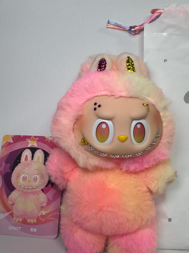 May include: A plush toy in shades of pink, yellow, and orange, resembling a bunny with a hood. The toy has a detailed face with large eyes and a small yellow nose. A card with a matching illustration is next to the toy. The word "LOYALTY" is printed on the card.