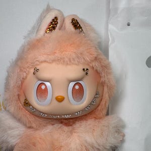 May include: A plush, peach-colored character with a bunny-like hood. The face has large, round eyes with gradient shading, a small nose, and a wide, toothy grin. The bunny ears are adorned with gold glitter. The character is soft and fluffy.