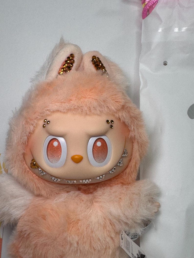 May include: A plush, peach-colored character with a bunny-like hood. The face has large, round eyes with gradient shading, a small nose, and a wide, toothy grin. The bunny ears are adorned with gold glitter. The character is soft and fluffy.