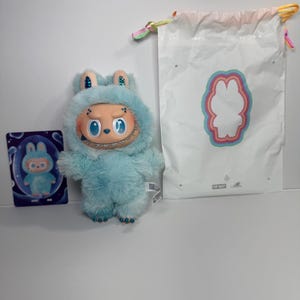 May include: A light blue plush toy with a smiling face and a furry hood, next to a matching card and a white drawstring bag with a bunny outline. The toy has a unique design and is likely a collectible item.
