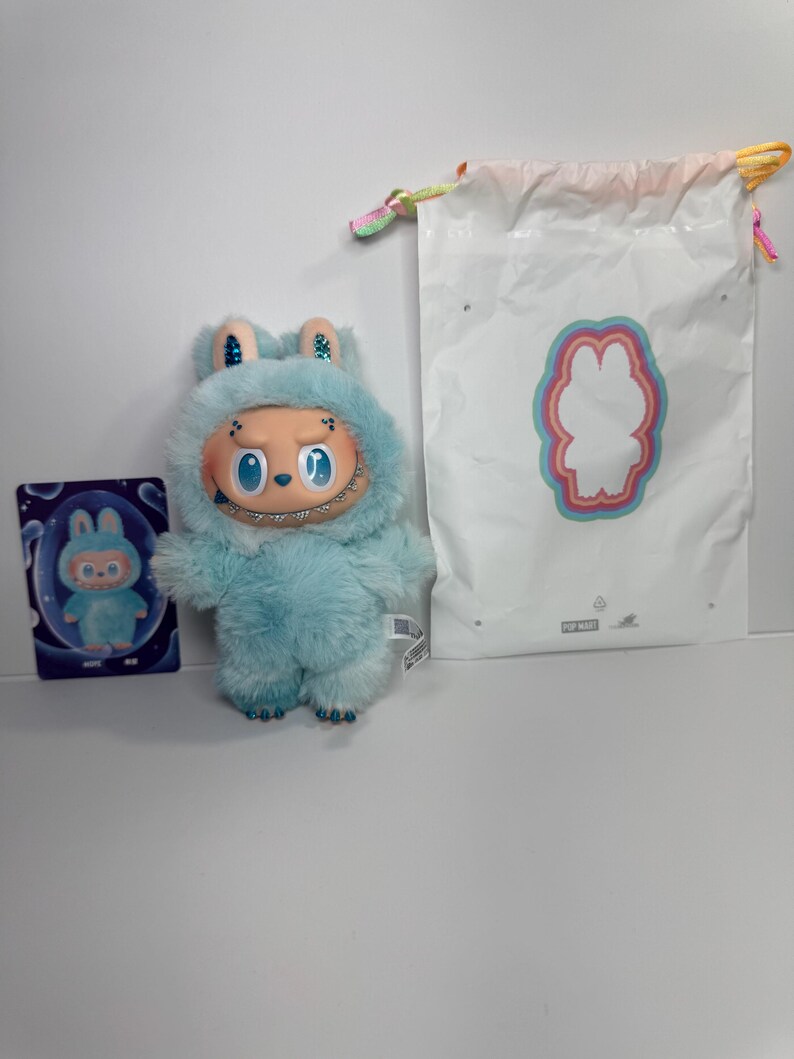 May include: A light blue plush toy with a smiling face and a furry hood, next to a matching card and a white drawstring bag with a bunny outline. The toy has a unique design and is likely a collectible item.