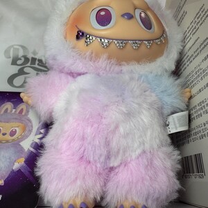 May include: A plush toy with a light tan face, purple eyes, and a jeweled smile. The toy is covered in fluffy, pastel-colored fur, with a white hood. The feet are adorned with jewels. A small card with a similar character is visible.