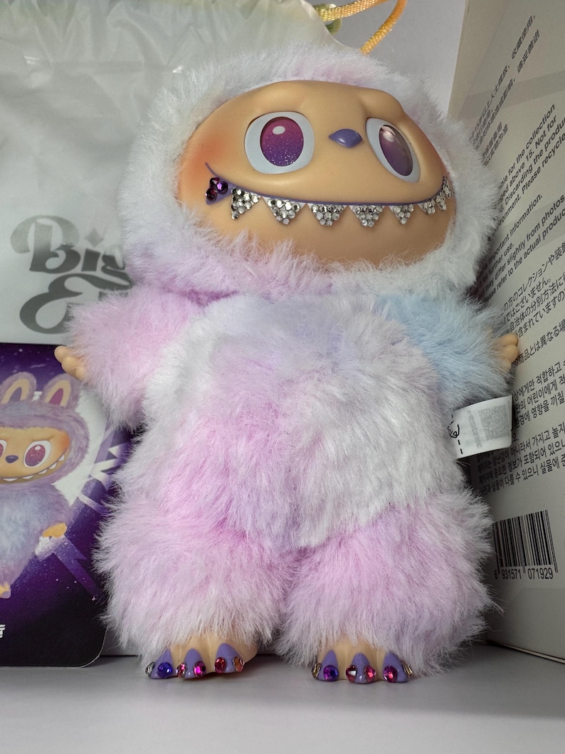 May include: A plush toy with a light tan face, purple eyes, and a jeweled smile. The toy is covered in fluffy, pastel-colored fur, with a white hood. The feet are adorned with jewels. A small card with a similar character is visible.