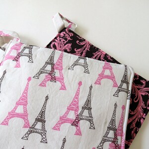 May include: Two pot holders with a white background. One features pink Eiffel Tower prints, the other has a black background with pink floral patterns. Both have white trim and hanging loops.