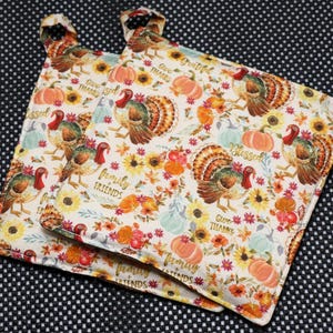 May include: Two Thanksgiving-themed pot holders with a white background. The pot holders feature a repeating pattern of turkeys, pumpkins, sunflowers, and fall foliage. The words "Give Thanks", "Blessed", and "Family Friends" are also visible.