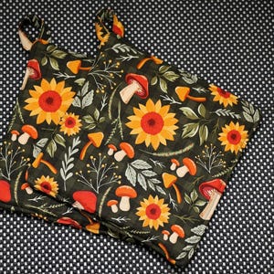 Könnte beinhalten: Zwei Topflappen mit dunklem Hintergrund, die ein Muster aus Sonnenblumen, Pilzen und Blättern aufweisen. Die Sonnenblumen sind gelb und orange, und die Pilze sind orange und rot. Die Topflappen haben Schlaufen zum Aufhängen.