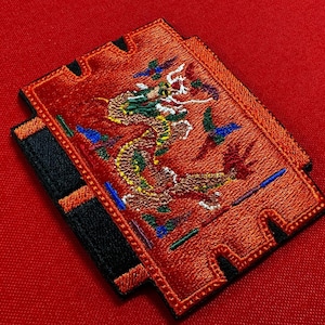 May include: Embroidered red patch with a dragon design in green, yellow, and brown. The patch has a black border and a distinctive shape with cut-out sections. The background is a solid red.