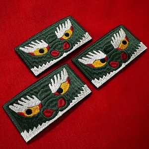 Pode incluir: Três patches bordados com um desenho de rosto de dragão. Os patches são retangulares com fundo verde escuro, olhos brancos e amarelos, detalhes vermelhos e costuras brancas na borda. São exibidos em uma superfície vermelha.