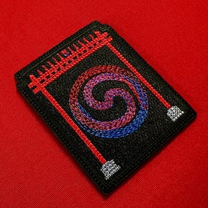 May include: Black embroidered patch featuring a spiral design in shades of red and blue. The patch has a red border with a geometric pattern at the top and two red lines with silver accents. The patch is rectangular.
