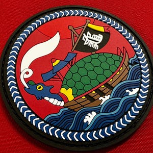 May include: A circular patch featuring a blue dragon-headed ship with a green turtle shell on its back, sailing on blue waves against a red background. The ship has a black sail with white Japanese text and a white plume of smoke rising from the dragon head.