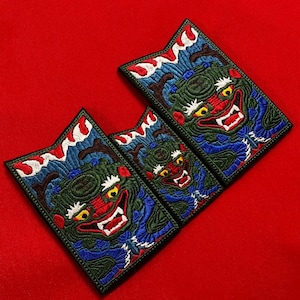 Pode incluir: Três patches bordados com um design de demônio japonês. Os patches apresentam um rosto vermelho com dentes brancos, detalhes verdes e azuis e um padrão de ondas na parte superior. O fundo é vermelho.