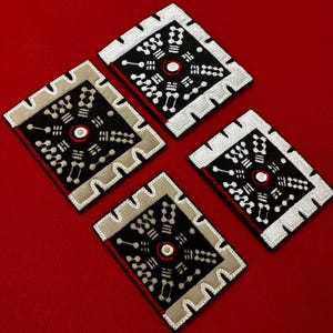May include: Four square embroidered patches with a geometric design. Each patch has a black centre with white and red accents, framed by a beige border and a white outer edge. The patches are arranged on a red background.