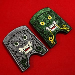 Peut inclure: Deux écussons brodés avec un motif de masque. L'un est gris avec des accents verts, l'autre est vert avec des accents jaunes. Les deux ont un visage souriant avec des dents pointues, sur fond rouge.