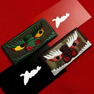 May include: Embroidered patches with detailed designs of stylized creatures in green, brown, and yellow, set against a red background. Each patch is rectangular with a textured appearance. The patches are accompanied by red, black, and white cards with a white logo.
