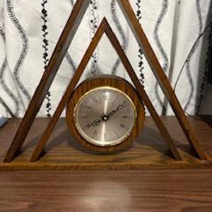 May include: A wooden clock with a round face and gold-coloured trim, set within a geometric, triangular wooden frame. The clock's face displays numbers and hands. The frame is supported by a rectangular base. The background features a white curtain with black vertical patterns.