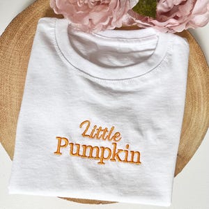 May include: White t-shirt with the embroidered text "Little Pumpkin" in orange. The shirt is folded and placed on a wooden tray, with pink flowers in the background. A simple, casual top.