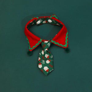 May include: A festive pet collar with a red and green design. The collar features a red body with green trim and a green tie with Santa Claus faces and the word "HO" printed on it. The collar has two green pom-poms.