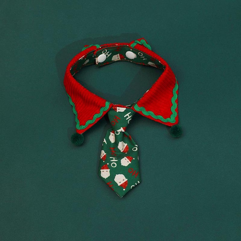 May include: A festive pet collar with a red and green design. The collar features a red body with green trim and a green tie with Santa Claus faces and the word "HO" printed on it. The collar has two green pom-poms.