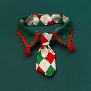 May include: A festive pet collar with a green, red, and white color scheme. The collar features a green ribbed design with red scalloped edges and a patterned tie. Two red pom-poms are attached to the collar, and it fastens with two white buttons.