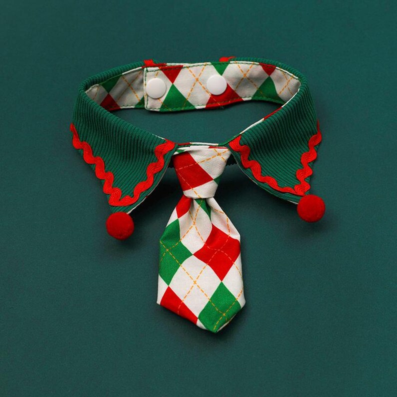 May include: A festive pet collar with a green, red, and white color scheme. The collar features a green ribbed design with red scalloped edges and a patterned tie. Two red pom-poms are attached to the collar, and it fastens with two white buttons.