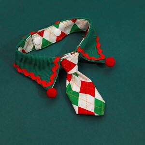 May include: A festive pet collar with a green corduroy base and a red and white argyle tie. The collar has a scalloped edge with red trim and small red pom-poms. Two white buttons secure the collar.