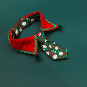 May include: A festive pet collar and tie set in red and green. The collar is red with a green scalloped edge and green pom-poms. The tie is green with Santa Claus faces and the word "HO" printed on it. The set is designed for the Christmas holiday.