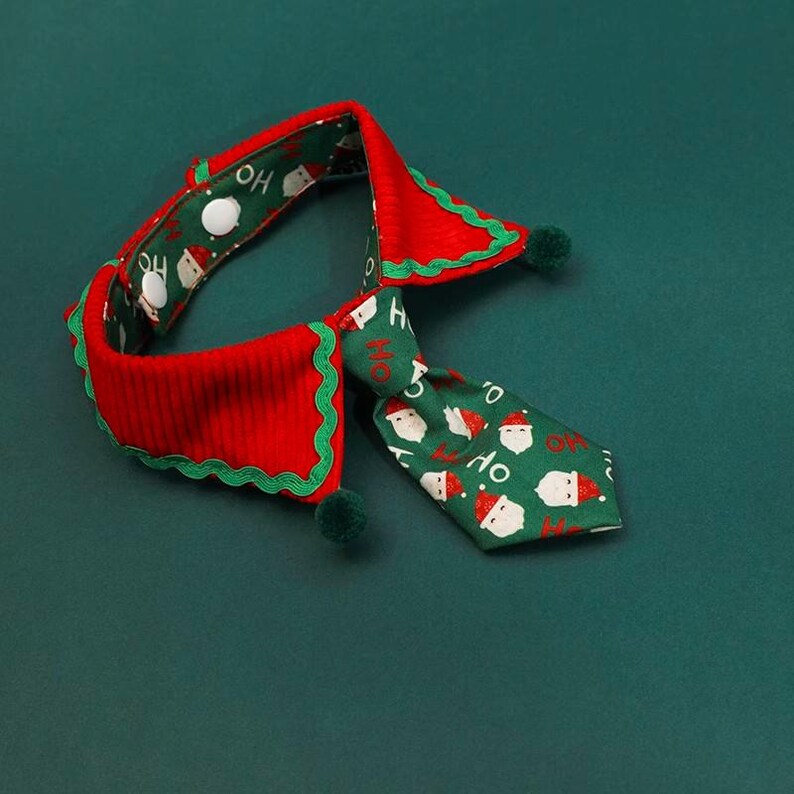 May include: A festive pet collar and tie set in red and green. The collar is red with a green scalloped edge and green pom-poms. The tie is green with Santa Claus faces and the word "HO" printed on it. The set is designed for the Christmas holiday.