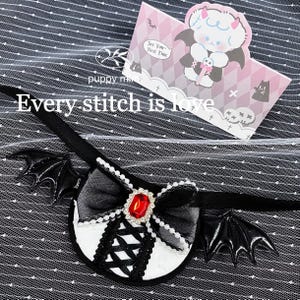 May include: Black and white gothic-style headband with bat wings. The headband features a black bow with a red jewel, and the text "Every stitch is love". A pink card with a cartoon character is in the background.