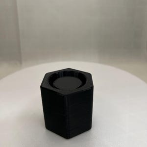 May include: A black, hexagonal-shaped container with a circular indentation at the top. The container is made of a solid, matte material and sits on a white surface. The design is simple and modern, suitable for various uses.