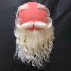 May include: A Santa Claus wig and beard set. The wig is a light pink color, and the beard and mustache are a long, curly, white color. The beard is attached to the wig. The set is designed for costume use.