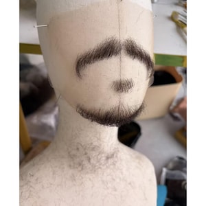 May include: A beige lace facial hair piece featuring a dark brown moustache, a small soul patch, and a chin beard. The hair is attached to a sheer, skin-toned mesh base that is secured to a mannequin head with silver pins for display and styling.