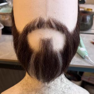 Peut inclure: Une barbe artificielle brun foncé et gris fixée sur une tête de mannequin beige. Les poils du visage sont disposés en un motif circulaire autour de la zone de la bouche, avec une moustache et une barbiche aux mèches de cheveux visibles.