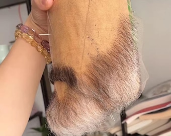 May include: A light brown lace front wig base with a gray and brown beard and mustache attached. The wig base is held up by a hand, showing the fine mesh and the realistic hair detailing. The wig appears to be for cosplay or costume use.