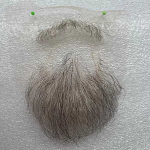 May include: A gray and black fake beard and mustache set. The beard is a full, rounded shape, and the mustache is a thin strip. The set is pinned to a white surface with two green pins. The hair appears to be synthetic.