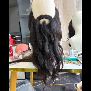 May include: A dark brown, wavy hairpiece styled as a beard and mustache, attached to a beige mannequin head. The facial hair features a defined mustache area and long, flowing wavy strands that extend downward from the chin area.
