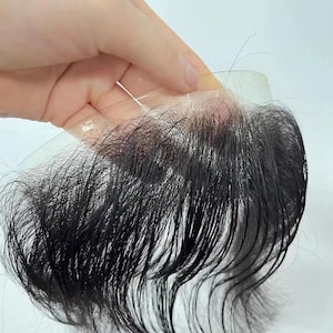 May include: A dark brown hairpiece with a transparent base is held up by a hand. The hairpiece appears to be a hair replacement system, likely a toupee or hair system, designed to blend with natural hair.