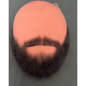 May include: A bald mannequin head with a dark brown beard and mustache. The beard is full and well-defined, with a natural appearance. The mannequin head is peach-colored, and the beard is attached to the head.