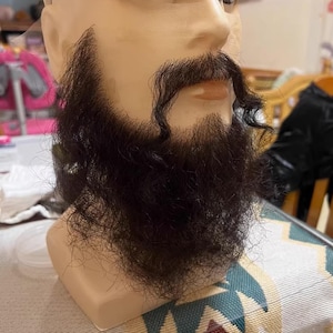 Op de afbeelding: Een donkerbruine baard en snor bevestigd aan een mannequinhoofd. De baard is vol en bedekt de kin en kaaklijn, met een licht gekrulde snor. Het mannequinhoofd heeft een lichte huidskleur.