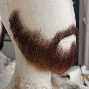 Peut inclure: Une barbe brune et auburn fixée sur une tête de mannequin. La barbe est coiffée avec une forme pleine, et les cheveux semblent être faits de fibres synthétiques. La tête de mannequin est recouverte d'un tissu clair, et des épingles sont insérées pour maintenir la barbe en place.