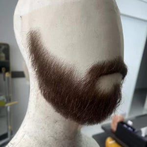 May include: A brown beard attached to a mannequin head. The beard is a medium brown colour and has a full, well-groomed appearance. The mannequin head is a light beige colour.