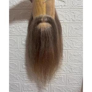 Op de afbeelding: Een lichtbruine baard bevestigd aan een houten vorm, gepresenteerd tegen een witte bakstenen muur. De baard heeft een natuurlijk, gelaagd uiterlijk met verschillende tinten bruin en blond.
