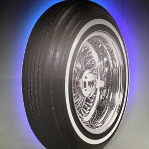 May include: A black and white image of a car tire with a chrome wire wheel and a white stripe. The tire is illuminated with a blue light around the edge, creating a striking visual effect. The image is a wall decoration.