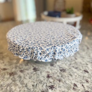 May include: A round, white and blue floral patterned fabric cover. The cover is draped over a round object, with the fabric gathered around the edges. The background is blurred, but appears to be a kitchen or dining area.