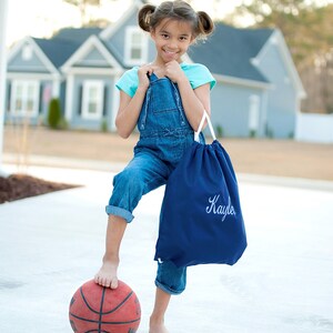 May include: A young girl wearing blue overalls and a turquoise shirt is standing on a basketball. She is holding a blue drawstring bag with the name "Kaylee" embroidered on it.