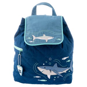May include: A blue quilted backpack with a shark design. The backpack has a flap with a smaller shark design and a zipper closure. The backpack has a small shark-shaped charm attached to the zipper.