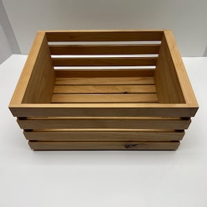 May include: A light brown wooden crate with a square shape. The crate is constructed with horizontal slats on the sides and bottom, and solid wood at the corners. The wood has a natural finish.