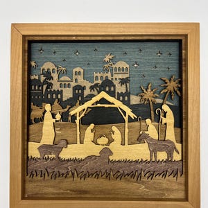 May include: A wooden nativity scene in a square frame. The layered design features a stable, figures, and a town silhouette against a blue sky with stars. The frame is a light brown color.