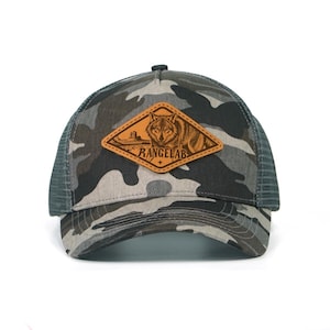 May include: Camouflage trucker hat with a mesh back and a curved brim. The front features a leather patch with a wolf illustration and the text "RANGELAB". The hat is a neutral color.